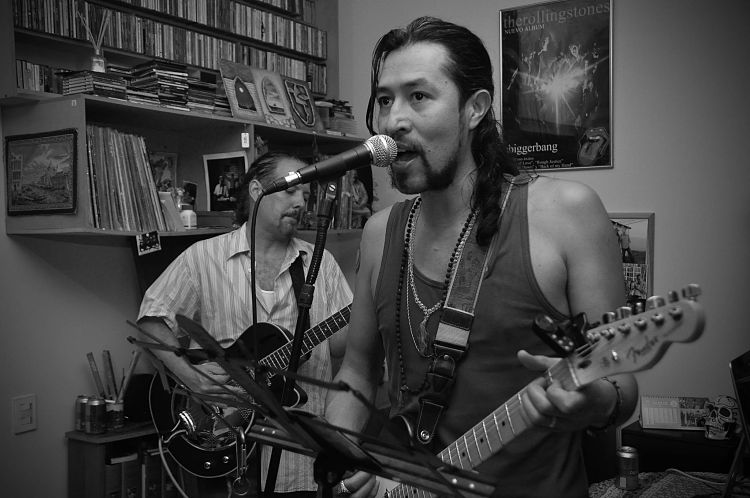 Un ensayo de Carlos Elliot Jr a blanco y negro tocando en primer plano con otro guitarrista de fondo