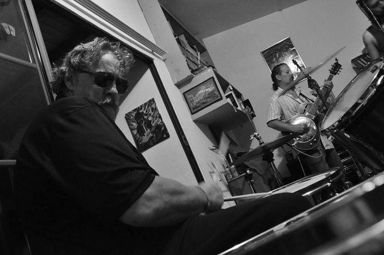 Un ensayo de Carlos Elliot Jr a blanco y negro tocando la bateria y la guitarra de fondo